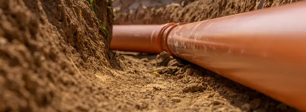 A large orange plastic pipe is laid in a narrow trench dug in the soil, with earth piled on both sides&mdash;typical of underground pipe installation for basement waterproofing in Montgomery & Chester County, PA.
