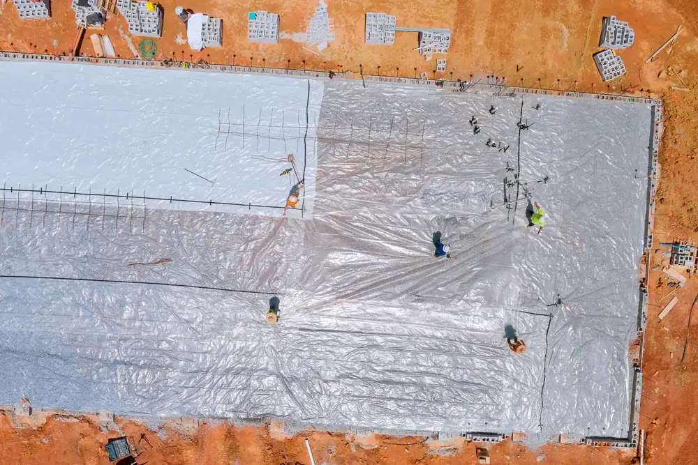 Aerial view of construction workers spreading large plastic sheets over a rectangular foundation area on reddish soil, a crucial step in basement waterproofing Montgomery & Chester County, PA, with scattered building materials around the site.