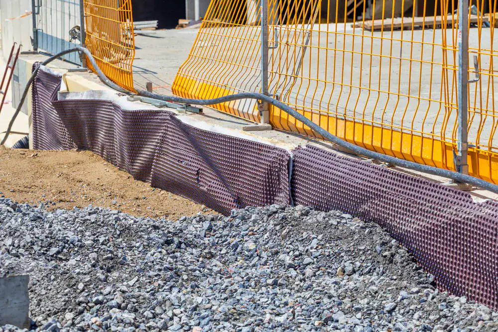 Construction site with gravel and soil in the foreground, a purple drainage membrane for basement waterproofing along a concrete wall, and yellow safety barriers and fencing in the background in Montgomery & Chester County, PA.