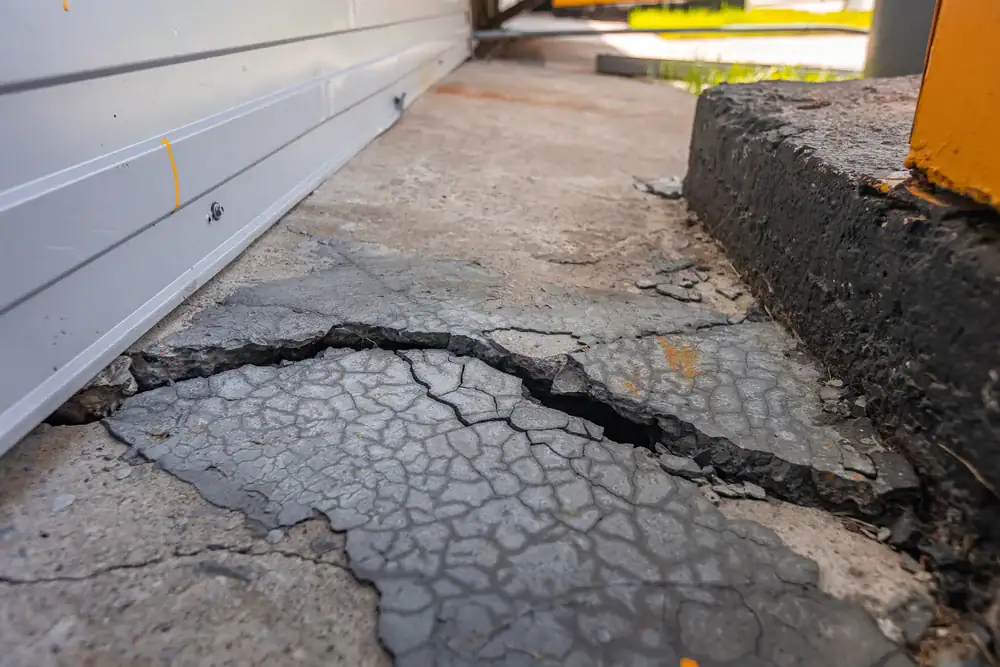 A large crack runs through the concrete sidewalk beside a building with a white metal wall and a yellow post, showing damaged and uneven pavement&mdash;an issue that may require Basement Waterproofing Montgomery & Chester County, PA.
