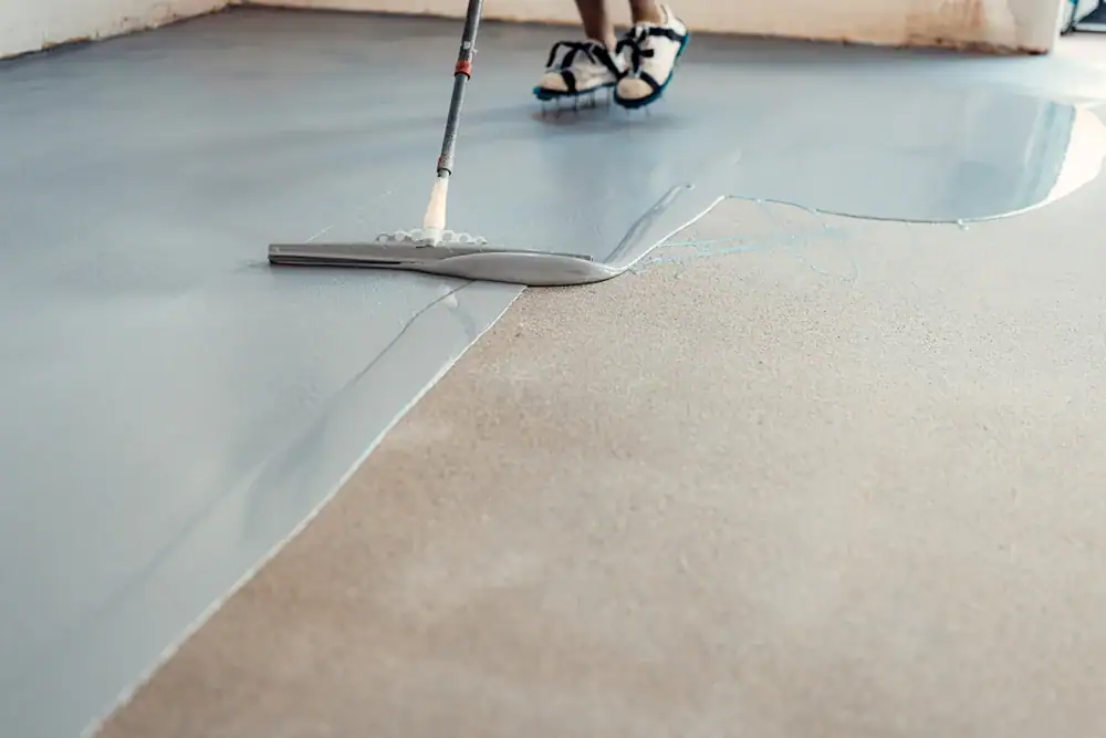 A person in spiked shoes is spreading self-leveling epoxy resin on a concrete floor with a large squeegee tool, part of a PA basement waterproofing project in Montgomery & Chester County.