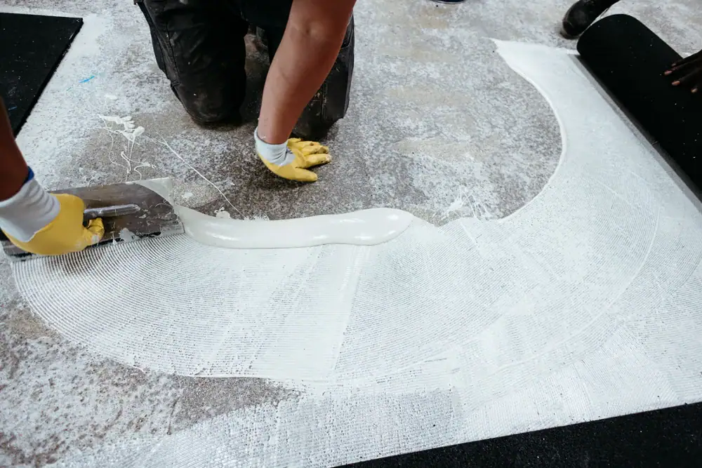 A person wearing yellow gloves spreads white adhesive with a trowel onto a textured floor, preparing the surface for new flooring installation.
