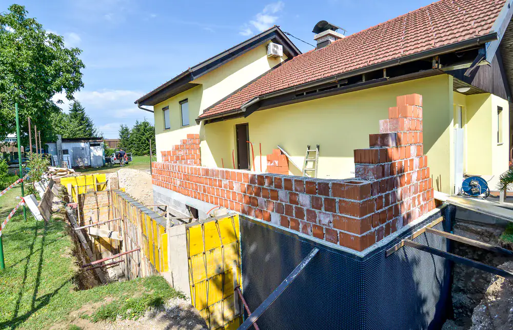 A house with yellow walls under renovation; new red brick walls are being constructed at the side, with foundation work and construction materials visible&mdash;ideal for those seeking basement waterproofing in Montgomery & Chester County, PA.