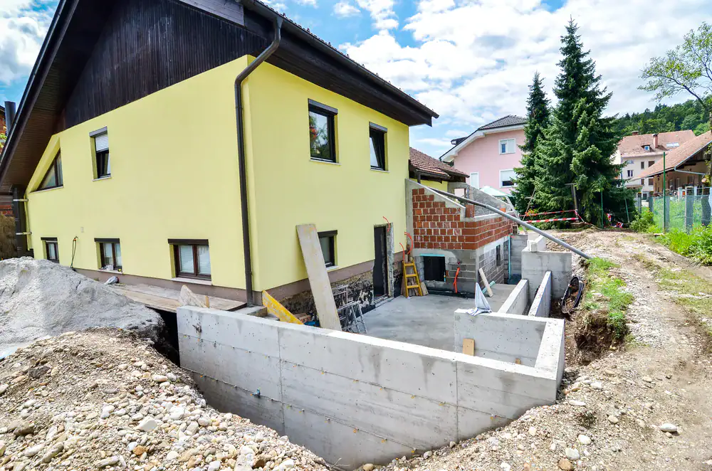 A yellow house with brown trim is undergoing construction, featuring exposed concrete walls and an unfinished extension&mdash;ideal for Basement Waterproofing Montgomery & Chester County, PA. Building materials and tools are visible, with piles of dirt surrounding the site on a sunny day.