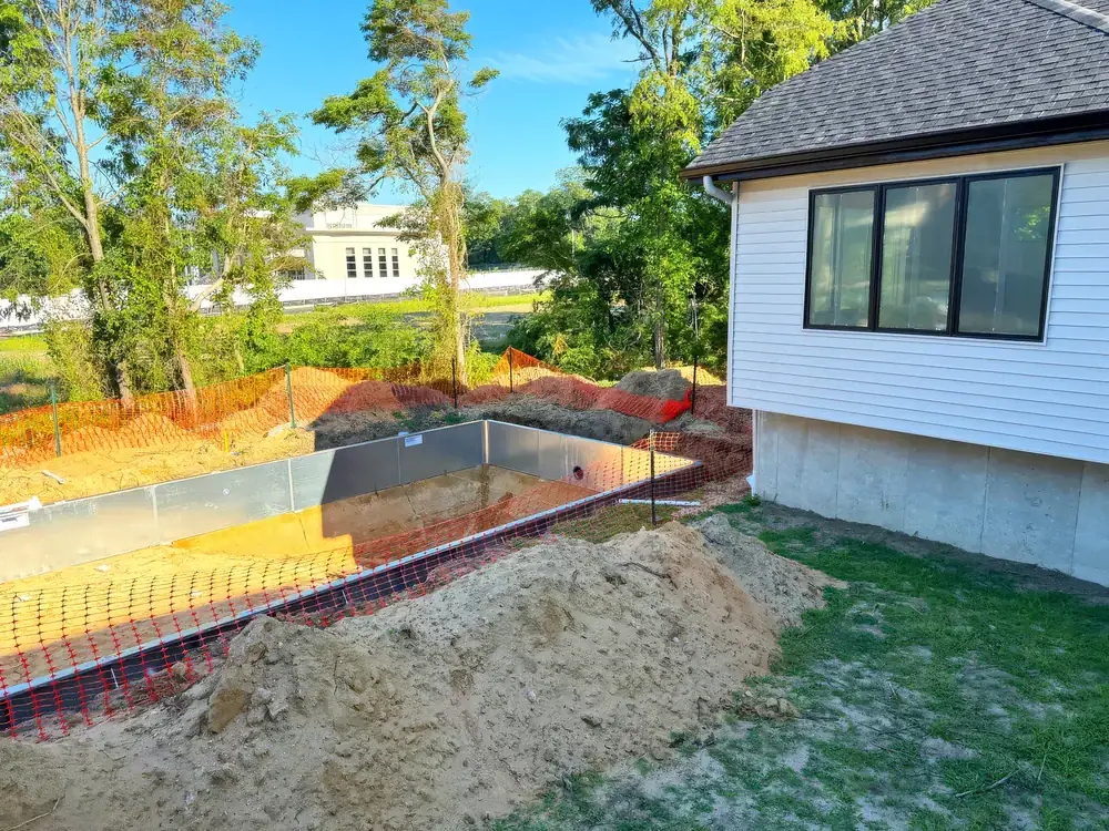 A backyard construction site with a rectangular hole for a swimming pool, surrounded by orange safety fencing and dirt piles, highlights the importance of basement waterproofing in Montgomery & Chester County, PA next to a white house with large windows and trees.