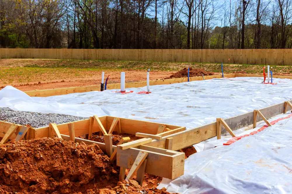 A house foundation under construction in PA, with wooden framing and plastic sheeting covering the ground. Exposed soil, plumbing pipes, and a background of trees hint at Basement Waterproofing Montgomery & Chester County underway.