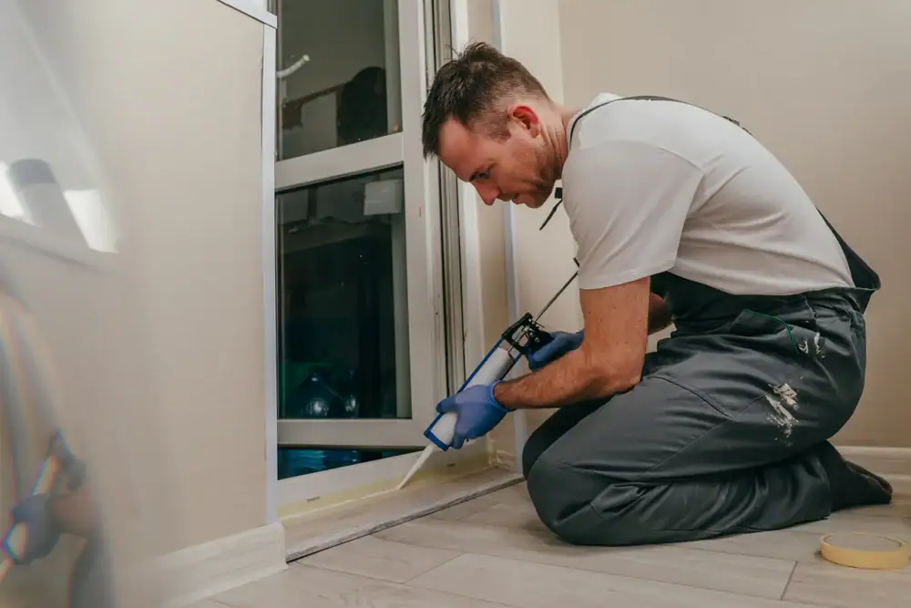A man in work overalls and blue gloves kneels on the floor and uses a caulking gun to seal the edge of a sliding glass door in a modern indoor space.