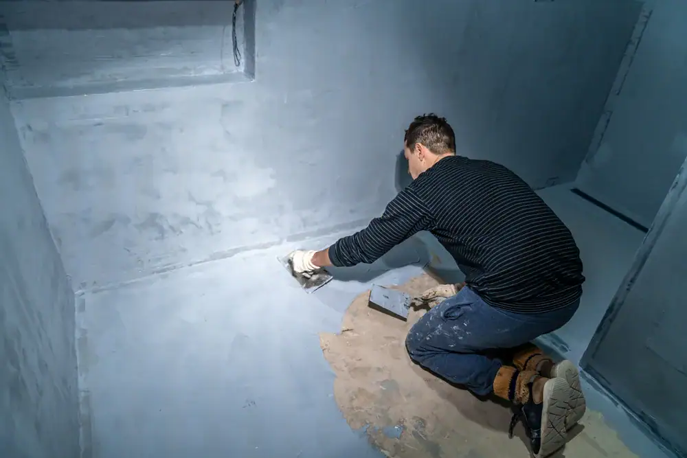A person wearing gloves and work clothes applies a gray waterproofing material to the floor and walls of a concrete room using a trowel.