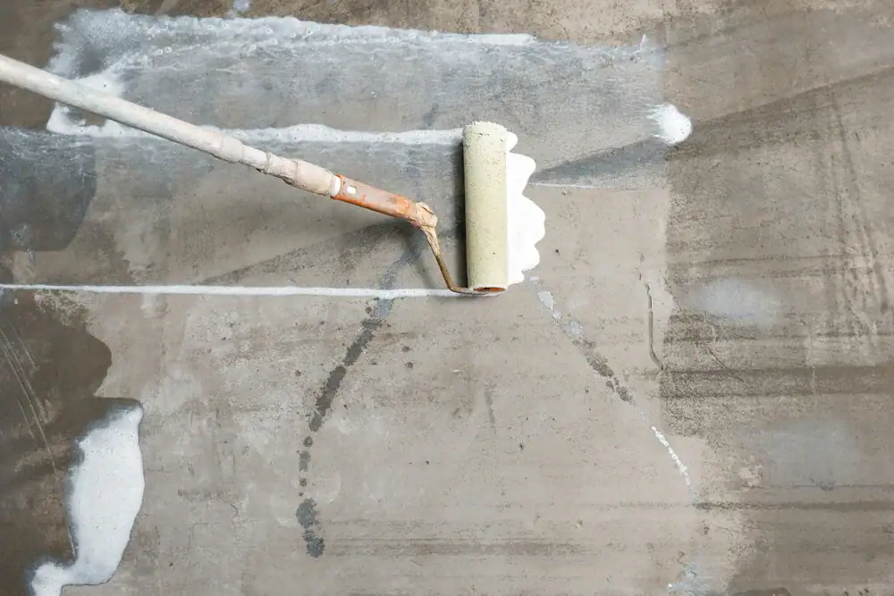 A paint roller is applying white primer or paint onto a bare concrete surface, creating wide, uneven strokes across the gray basement floor&mdash;ideal for PA basement waterproofing in Montgomery & Chester County.