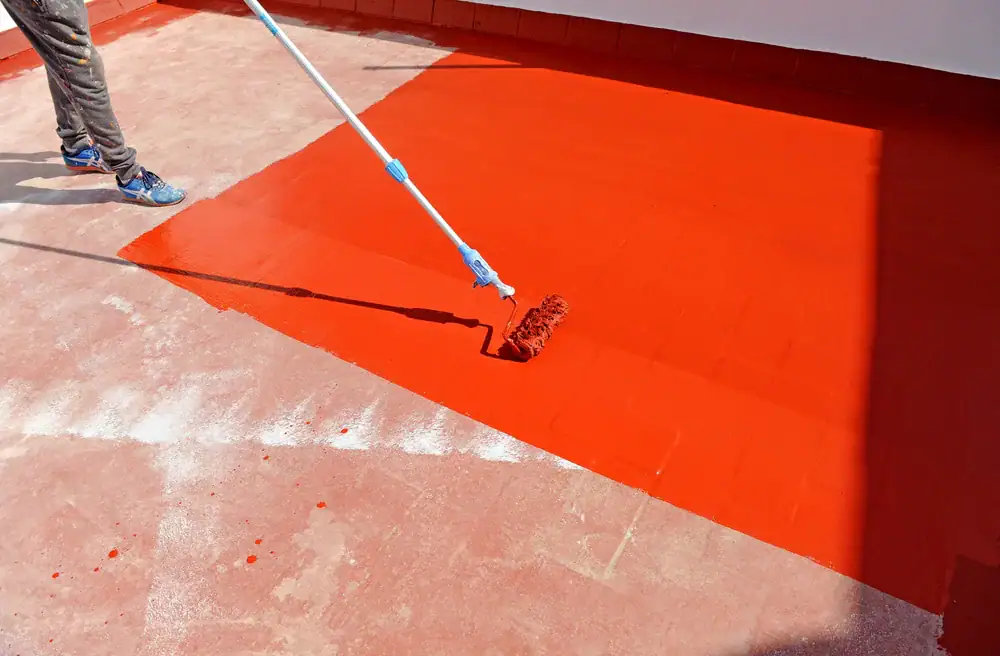 A person uses a long-handled paint roller to apply bright red paint onto a large cement surface, ideal for basement waterproofing in Montgomery & Chester County, PA, with part of the area already painted and part still unpainted.