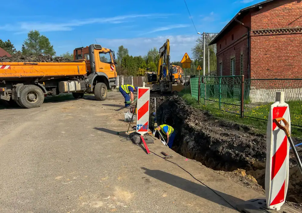 Construction workers are digging a trench along a road near a brick building for PA basement waterproofing in Montgomery & Chester County, with a truck and excavator nearby. Traffic barriers and warning signs secure the work zone under a clear sky.