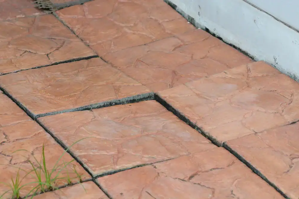 Several reddish-brown concrete tiles are unevenly placed outdoors, with some noticeably raised. Small gaps and visible grass grow between the tiles, a common issue that can signal drainage problems&mdash;consider Basement Waterproofing Montgomery & Chester County, PA.