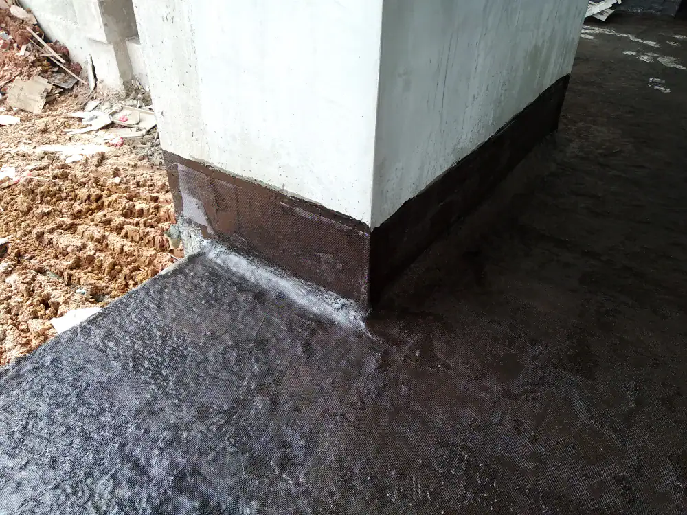 A building corner with walls and floor coated in dark waterproofing material, showcasing professional basement waterproofing Montgomery & Chester County, PA, next to a patch of exposed soil and construction debris.